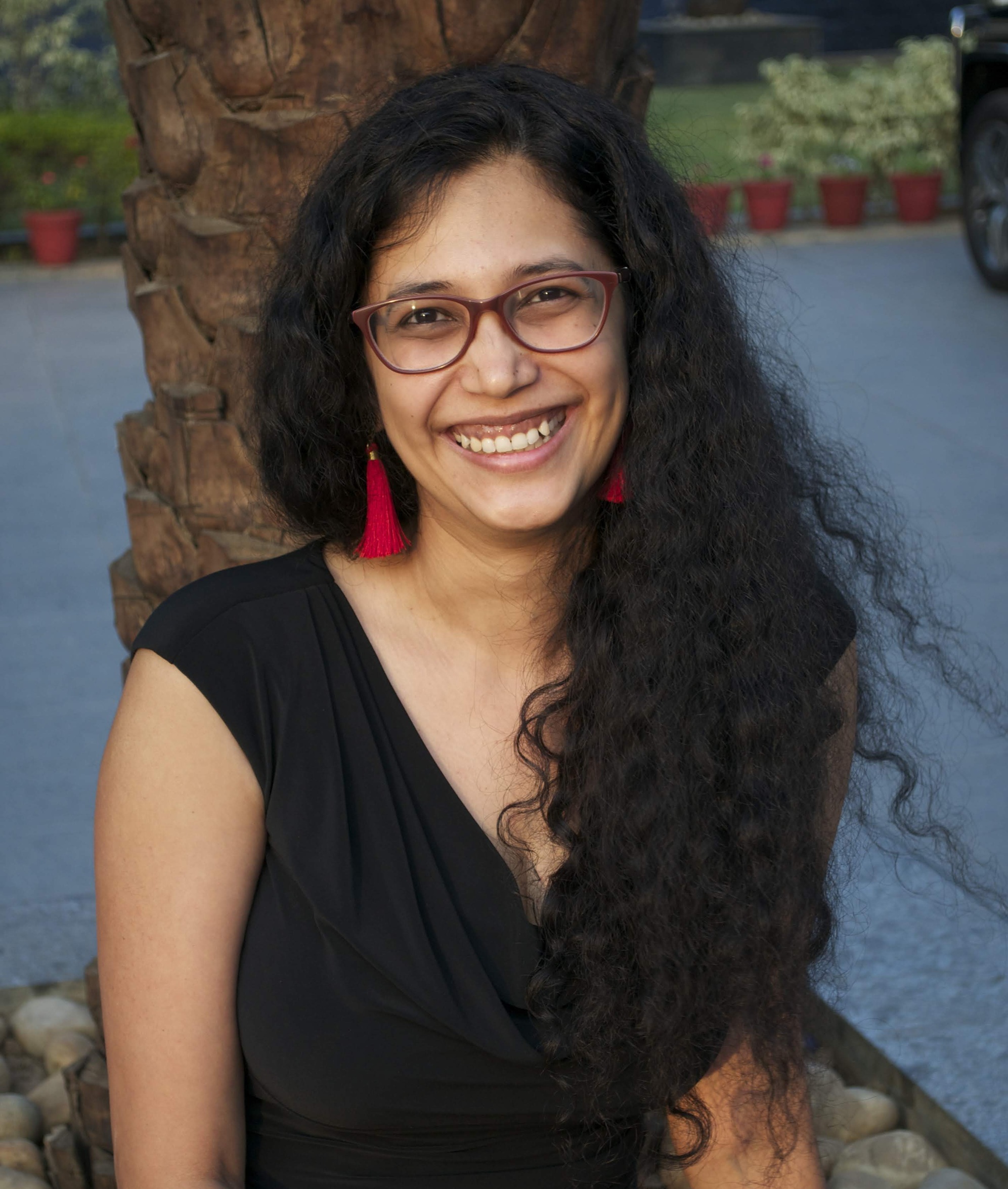 Gunjan Veda sitzt vor einer Palme, die in einem Topf auf einem Asphaltplatz steht. Sie trägt eine Brille, Ohrringe mit roten Fäden dran und ein schwarzes Kleid.