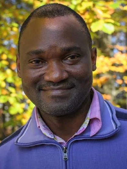 Emanuel Mbolela lächelt vor gelb-grünlich leuchtenden Laubbäumen in die Kamera. Er trägt ein hellviolettes Hemd und ein Pulli mit Kragen.